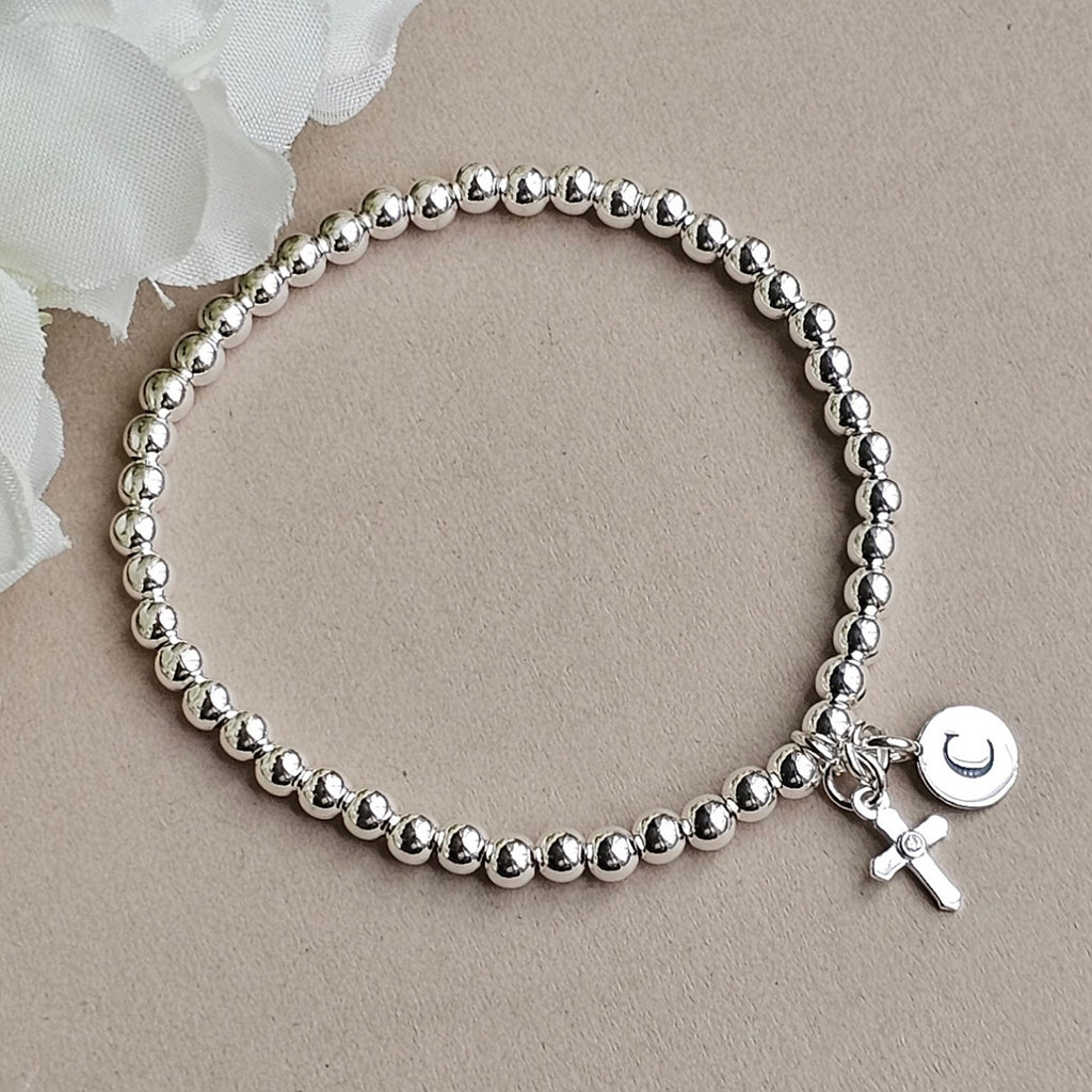 A silver beaded bracelet with a small charm featuring a cross and a round disc engraved with a letter 'C', displayed on a neutral beige background with a white flower in the corner.