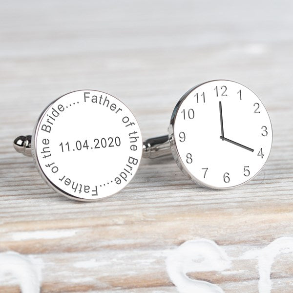 A pair of round silver cufflinks one has a face of a clock showing time and the other engraved with 'Father of the Bride' and a date displayed on a light wood background.