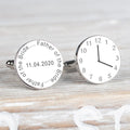 A pair of round silver cufflinks one has a face of a clock showing time and the other engraved with 'Father of the Bride' and a date displayed on a light wood background.