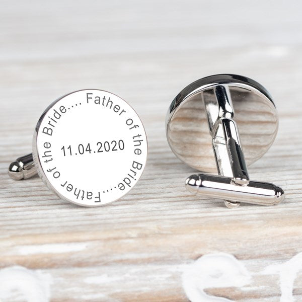 A pair of round silver cufflinks on a light wood surface. One cufflink is shown from the front with engraved text reading 'Father of the Bride' and a date.
