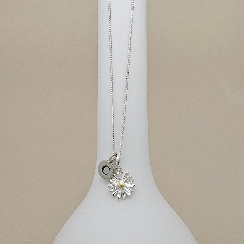 A silver necklace with a daisy flower pendant and a small round initial 'C' charm, displayed on a white neck form against a beige background.