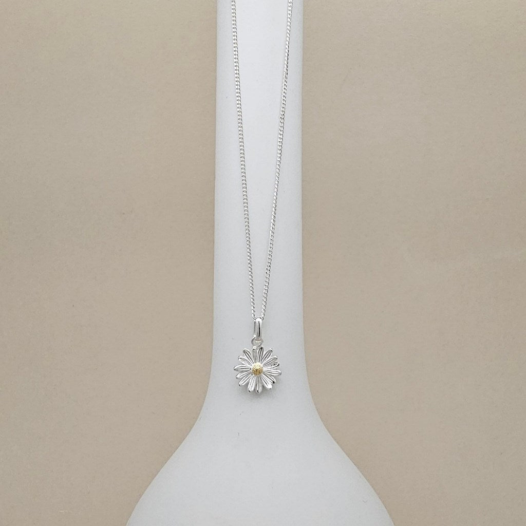 A silver daisy pendant necklace with a small yellow center, displayed on a white vase against a beige background.