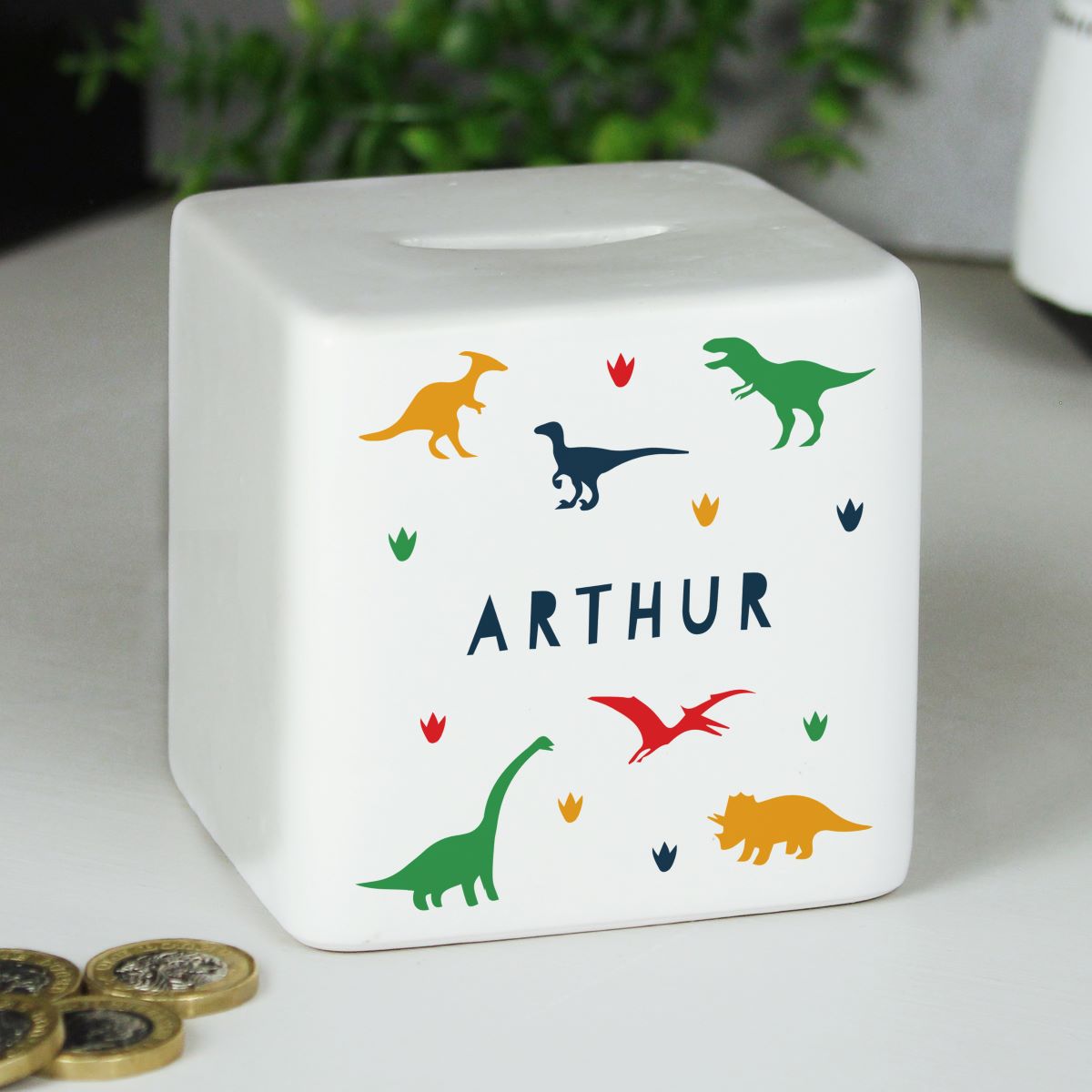 A white cube-shaped money box with dinosaur illustrations and the name ARTHUR printed on the front, photographed on a tabletop with coins and blurred greenery in the background.