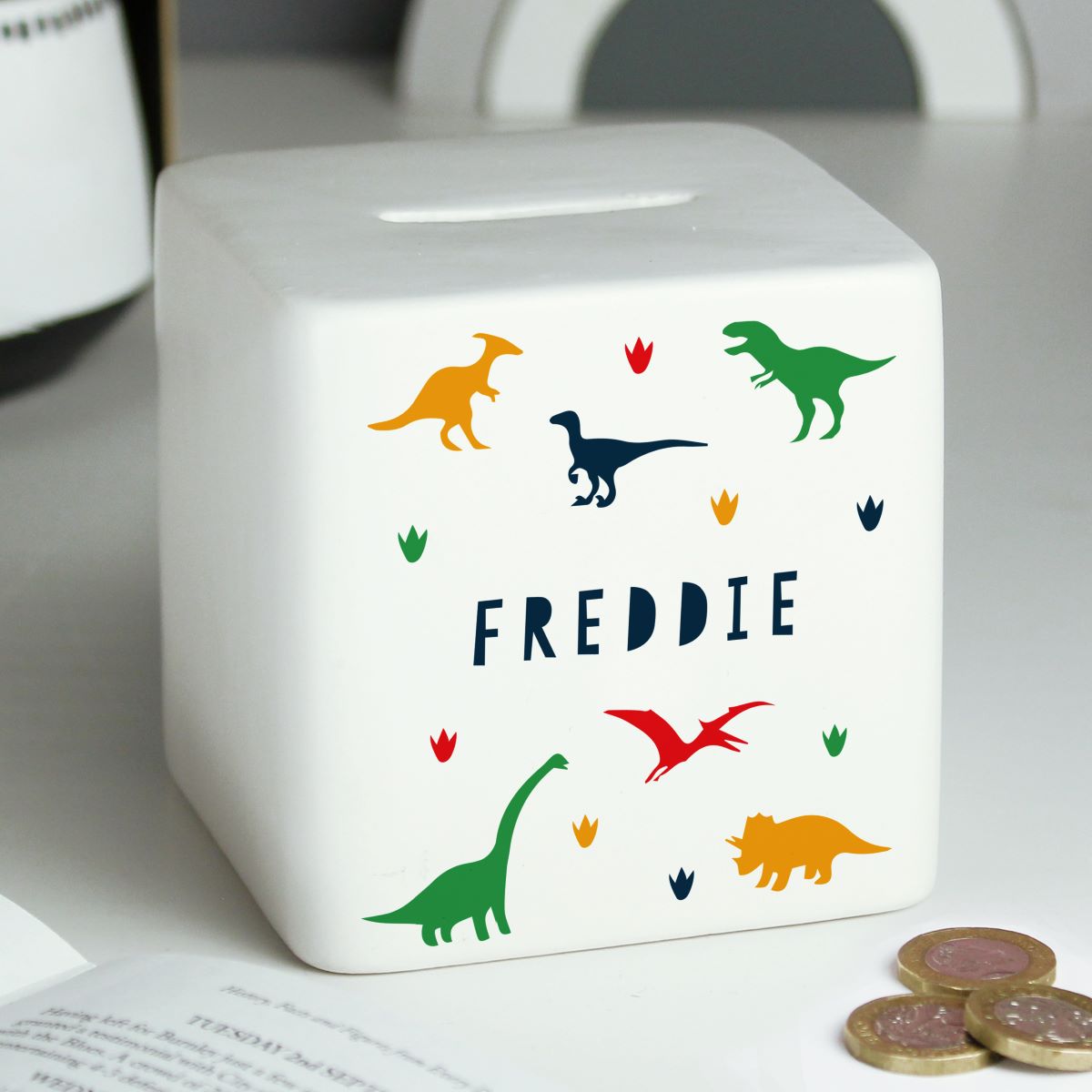 White cube-shaped money box with colorful dinosaur illustrations and the name 'Freddie' on a light gray surface.