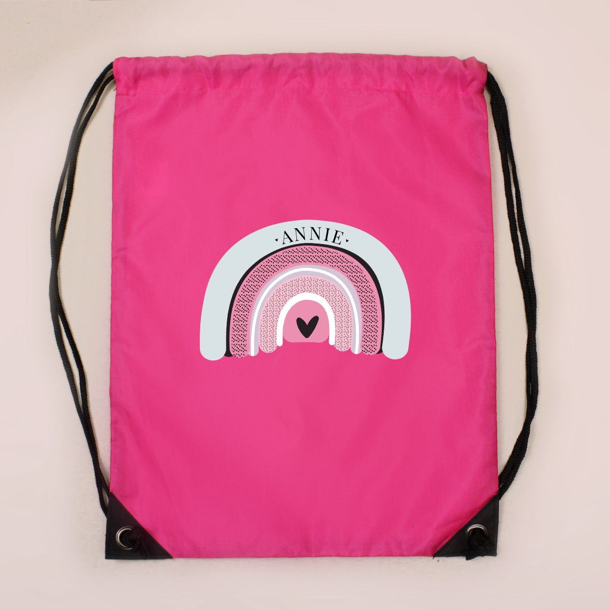 A bright pink drawstring bag with black cords and a central rainbow graphic that reads 'ANNIE' with a small heart in the middle, shown against a light beige background.