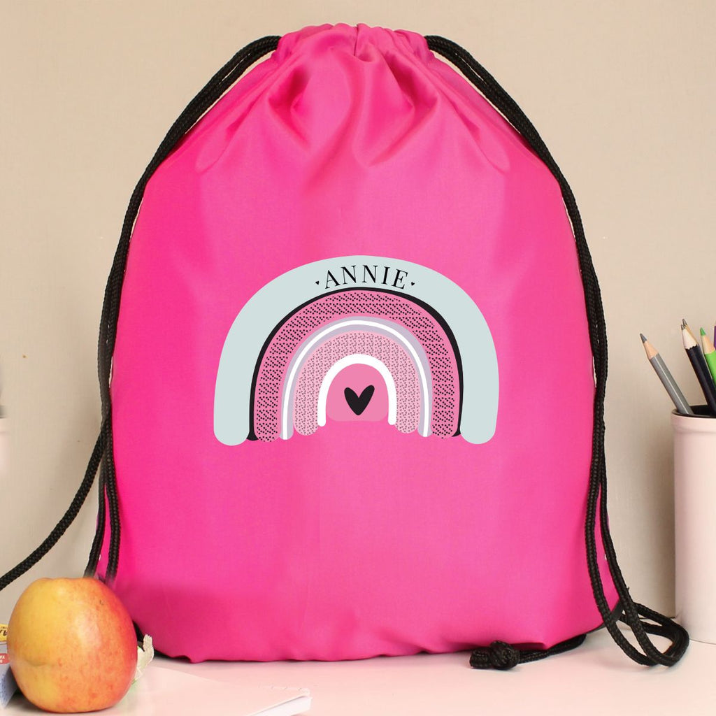 A bright pink drawstring gym bag with black cords, featuring a custom printed rainbow design with the name 'ANNIE' and a small heart. The bag is displayed on a light-colored surface next to an apple and a pencil cup.