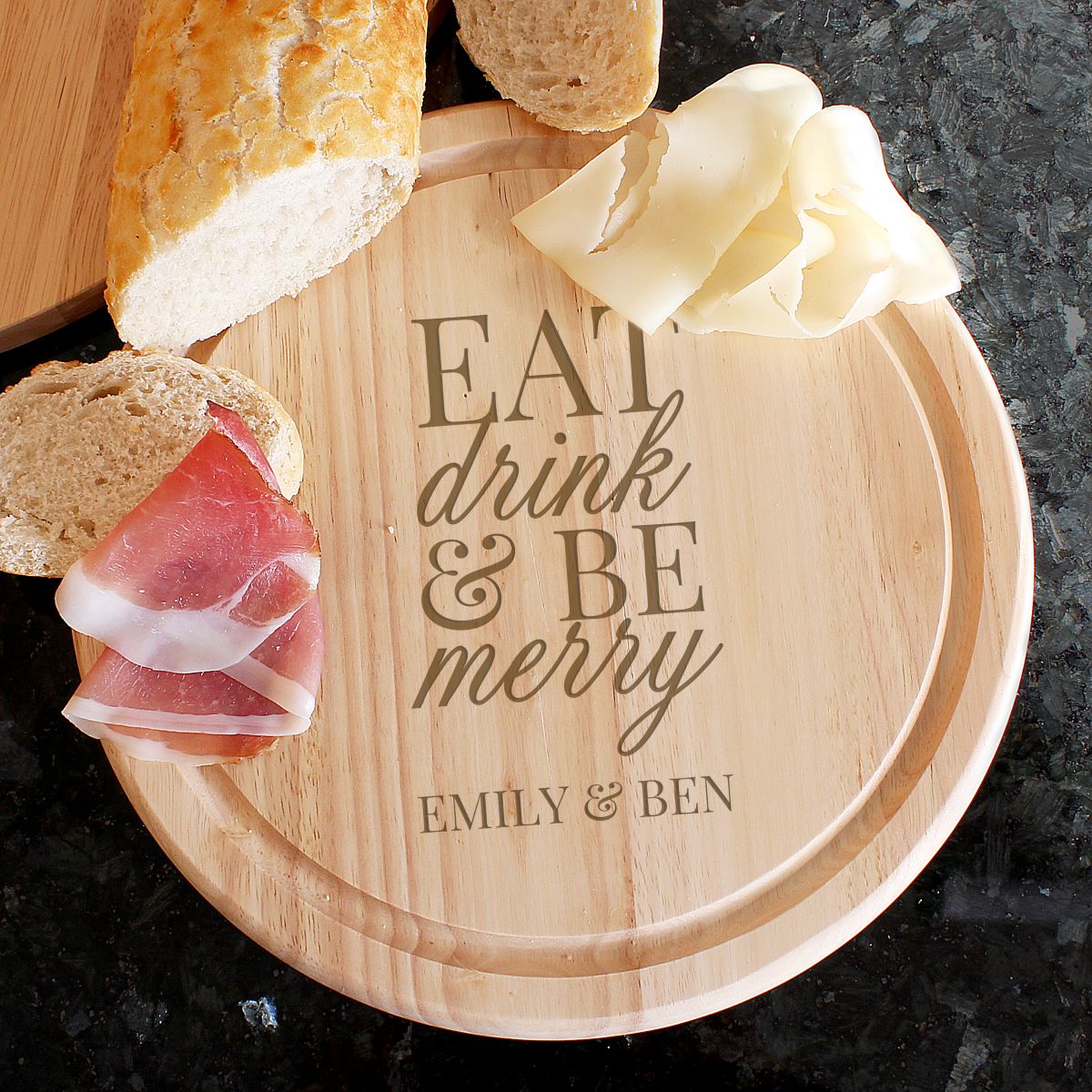 Wooden cutting board with 'Eat drink & be merry' engraving, bread, prosciutto, and cheese on a dark surface.