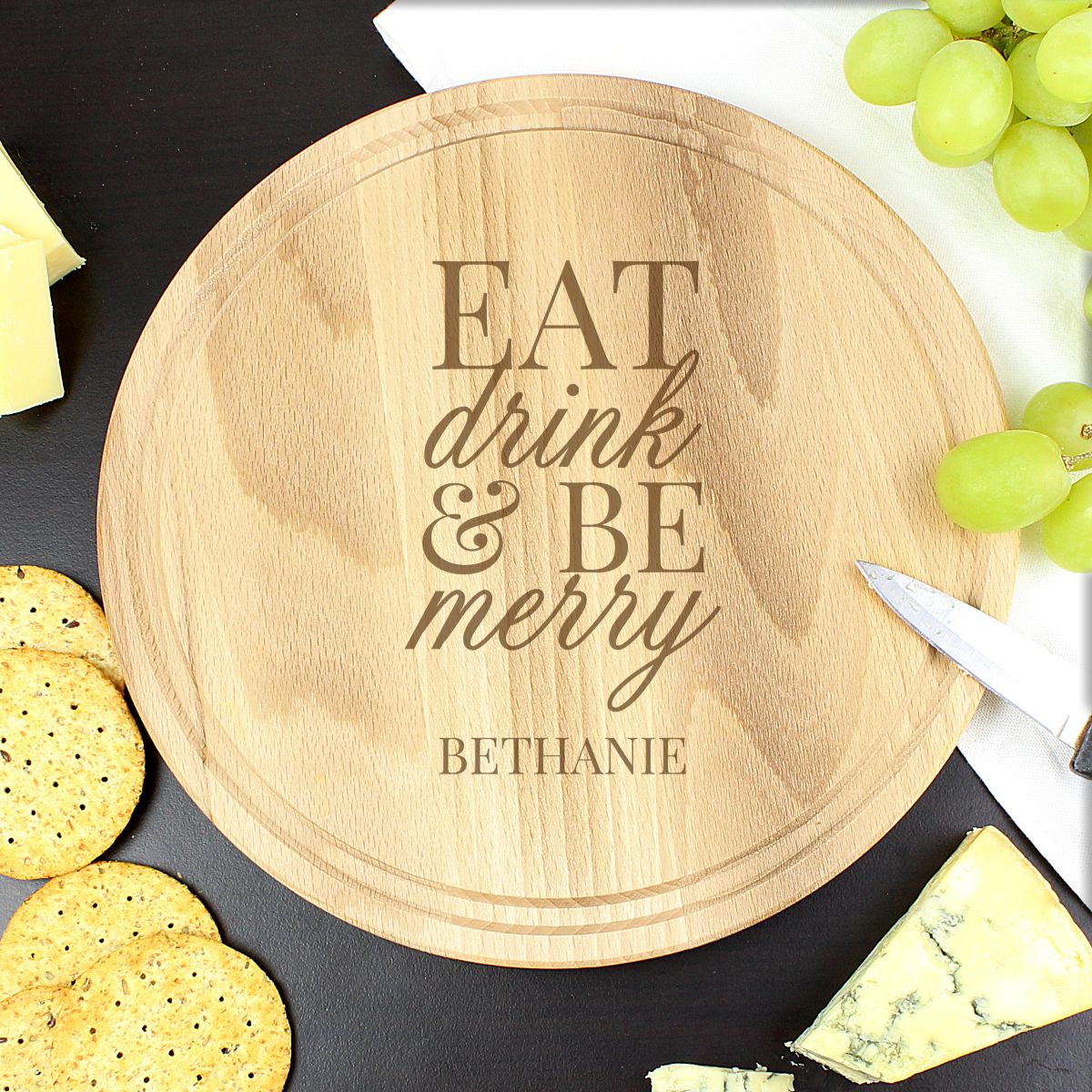 Wooden cutting board with 'Eat, drink & be merry' engraving on a table with cheese, crackers, and grapes.