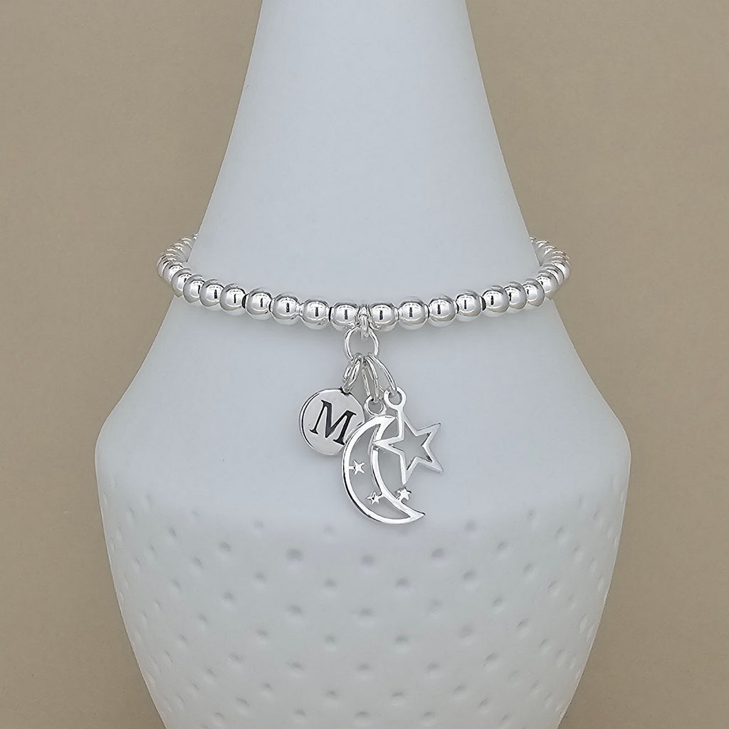 A silver beaded bracelet with a crescent moon, star, and initial 'M' charm, displayed on a white textured vase against a beige background.