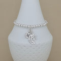 A silver beaded bracelet with a crescent moon, star, and initial 'M' charm, displayed on a white textured vase against a beige background.
