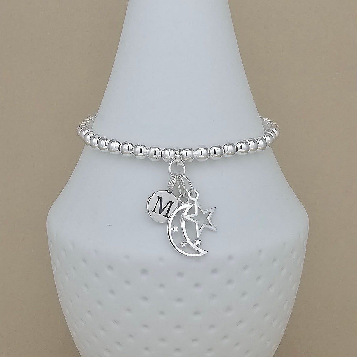 A silver beaded bracelet with a crescent moon, star, and initial 'M' charm, displayed on a white textured vase against a beige background.