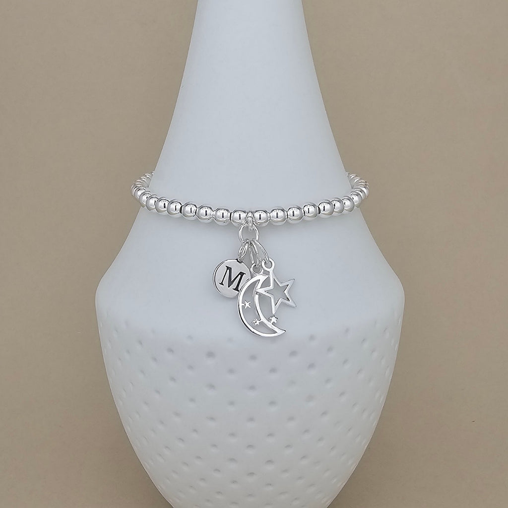 A silver beaded bracelet with dangling charms, including a crescent moon, a star, and a round initial 'M' charm, displayed on a white cone stand against a beige background.