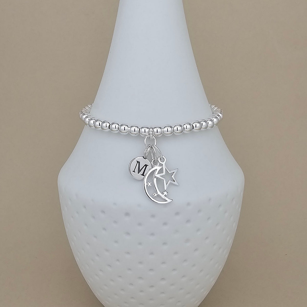 A silver beaded bracelet with dangling charms, including a crescent moon, a star, and a round initial 'M' charm, displayed on a white cone stand against a beige background.