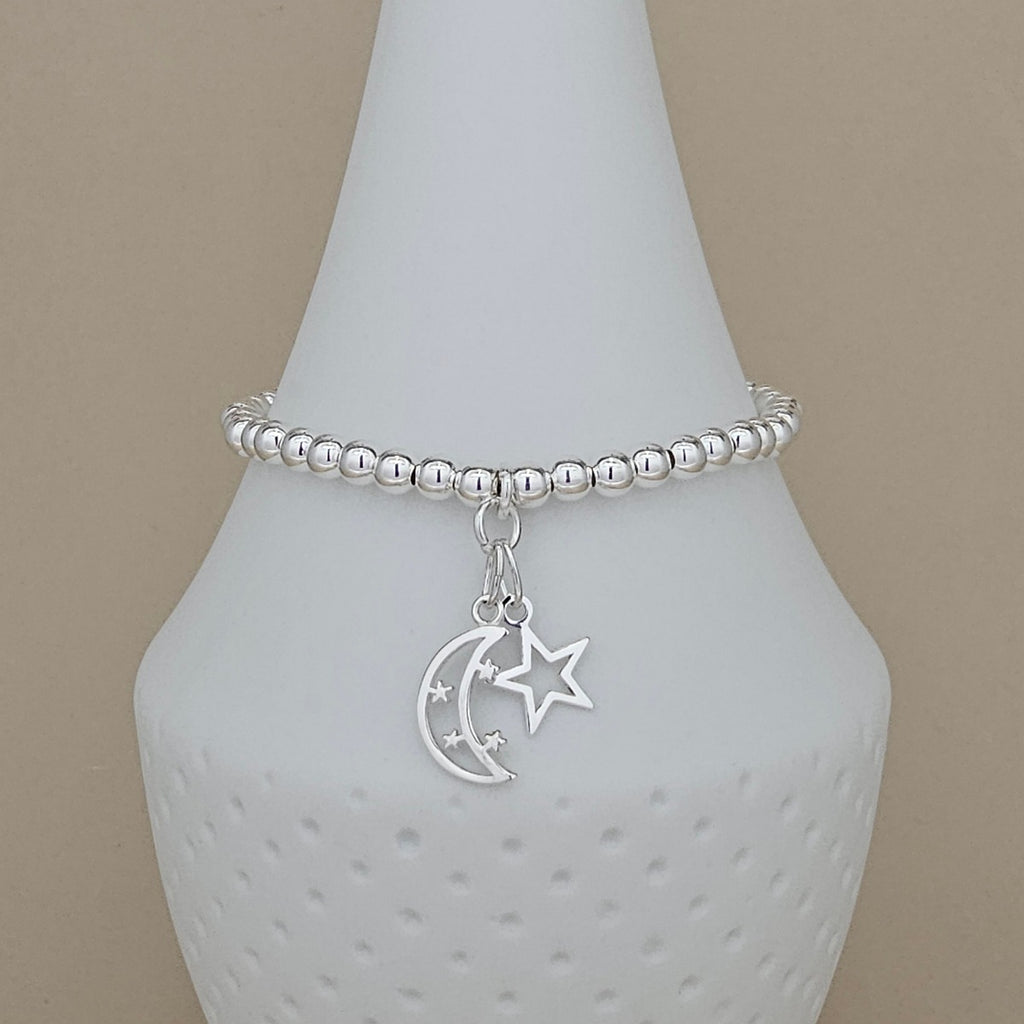 A silver beaded bracelet with a dangling crescent moon and star charm, displayed on a white ceramic jewelry stand against a beige background.