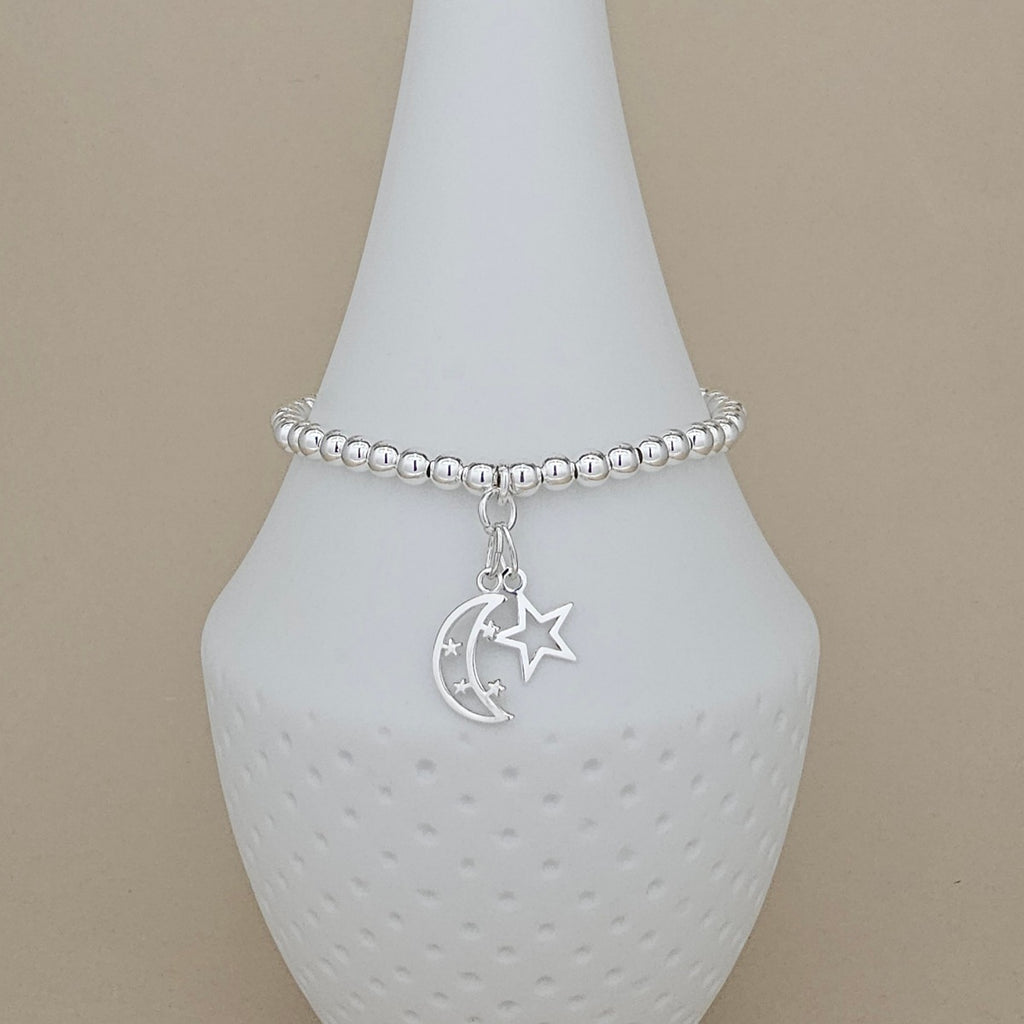 A silver beaded bracelet with a crescent moon and star charm, displayed on a white ceramic cone stand against a beige background.