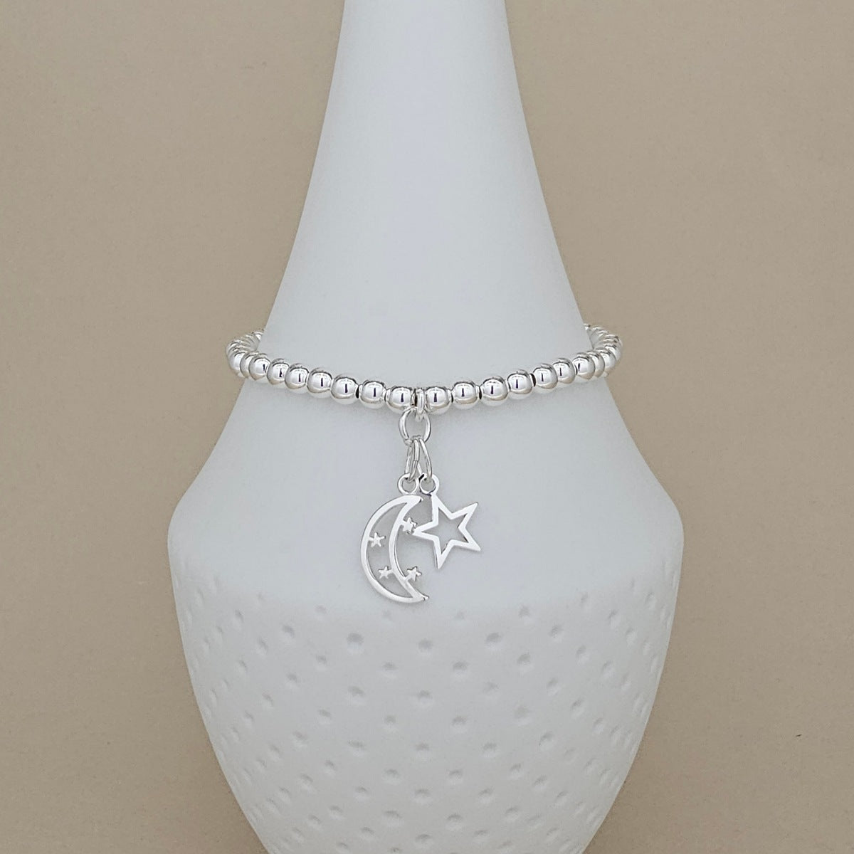 A silver beaded bracelet with a crescent moon and star charm, displayed on a white ceramic cone stand against a beige background.