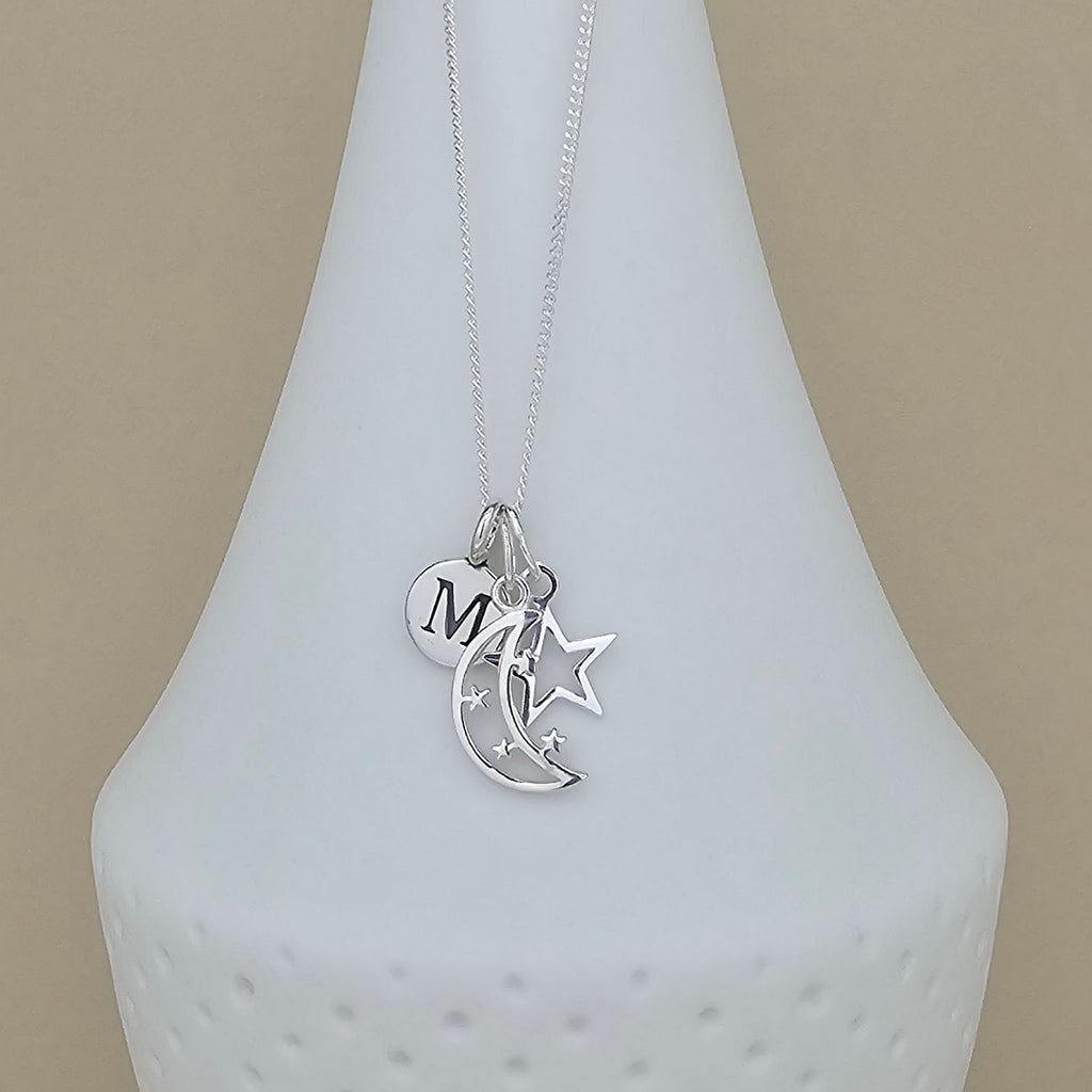 A silver necklace with a delicate chain and three charms: a heart engraved with the letter 'M', a crescent moon, and a star, displayed on a white ceramic vase against a beige background.
