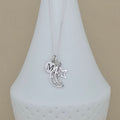 A silver necklace with a delicate chain and three charms: a heart engraved with the letter 'M', a crescent moon, and a star, displayed on a white ceramic vase against a beige background.