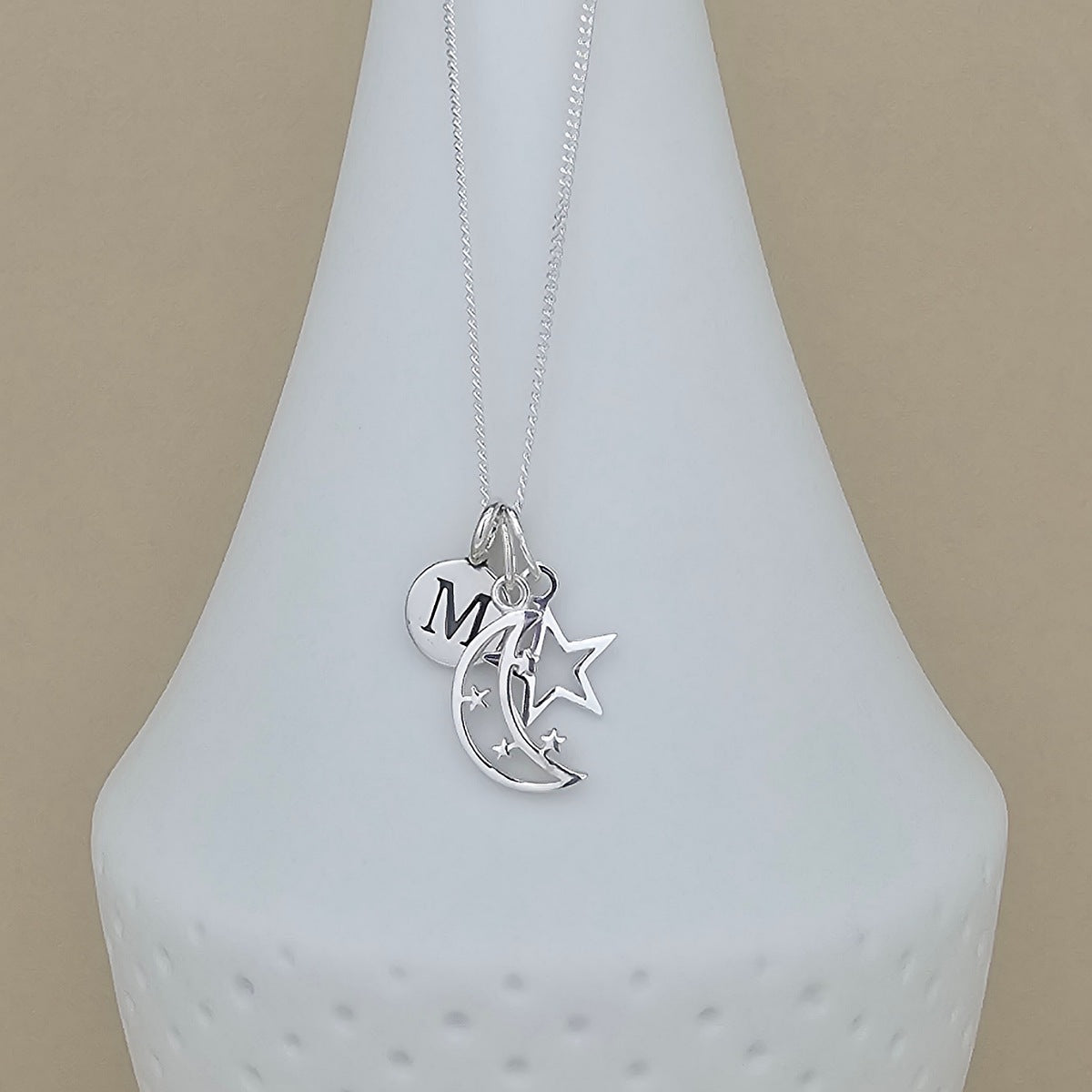 A silver necklace with a delicate chain and three charms: a heart engraved with the letter 'M', a crescent moon, and a star, displayed on a white ceramic vase against a beige background.