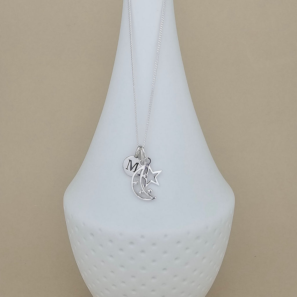 A silver necklace with a crescent moon and star pendant, featuring a small heart charm engraved with the letter 'M', displayed on a white ceramic vase against a beige background.
