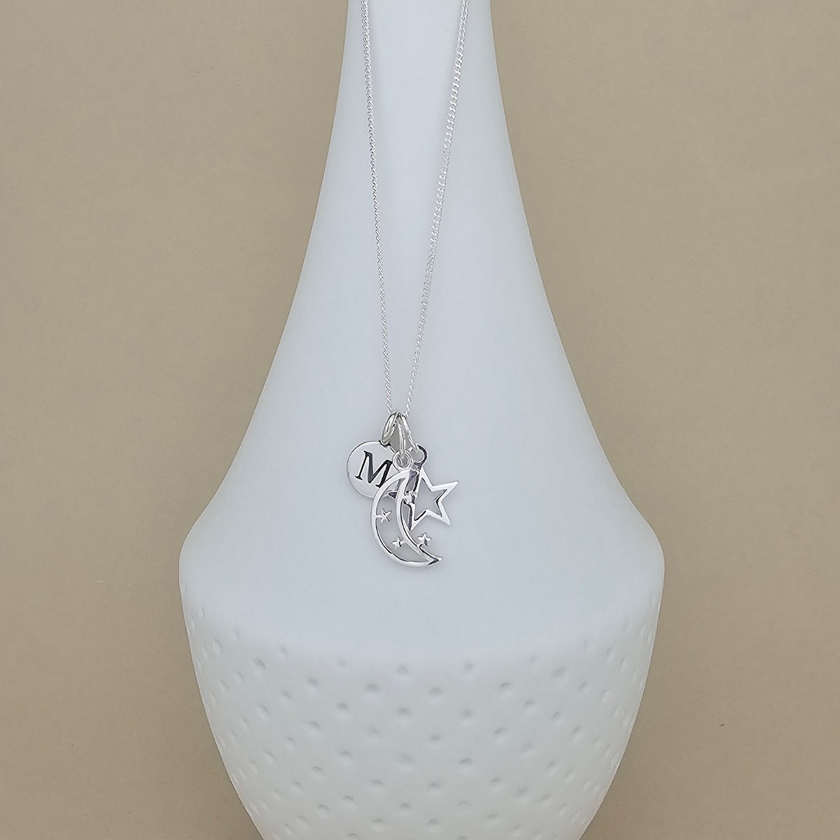 A silver necklace with a crescent moon and star pendant, featuring a small heart charm engraved with the letter 'M', displayed on a white ceramic vase against a beige background.