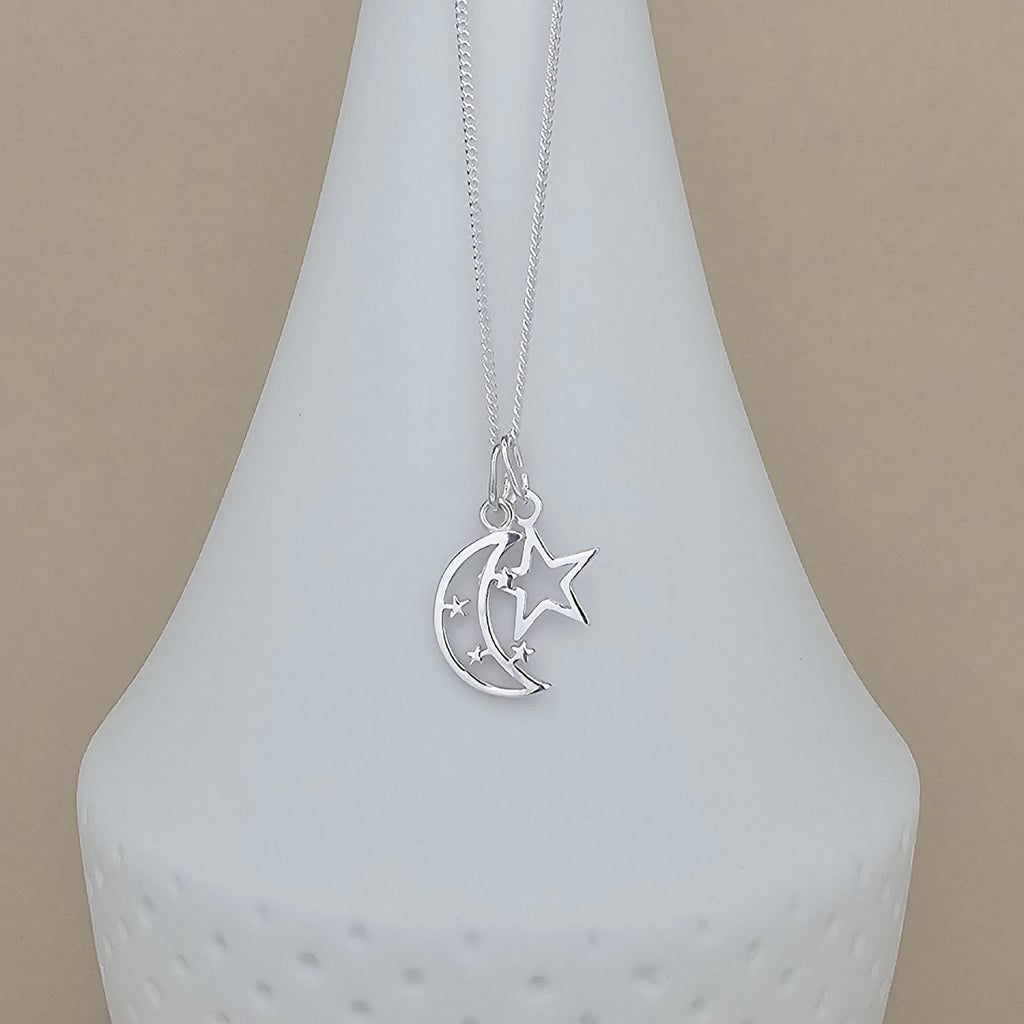 A silver necklace with a crescent moon and star pendant, displayed on a white ceramic vase against a beige background.