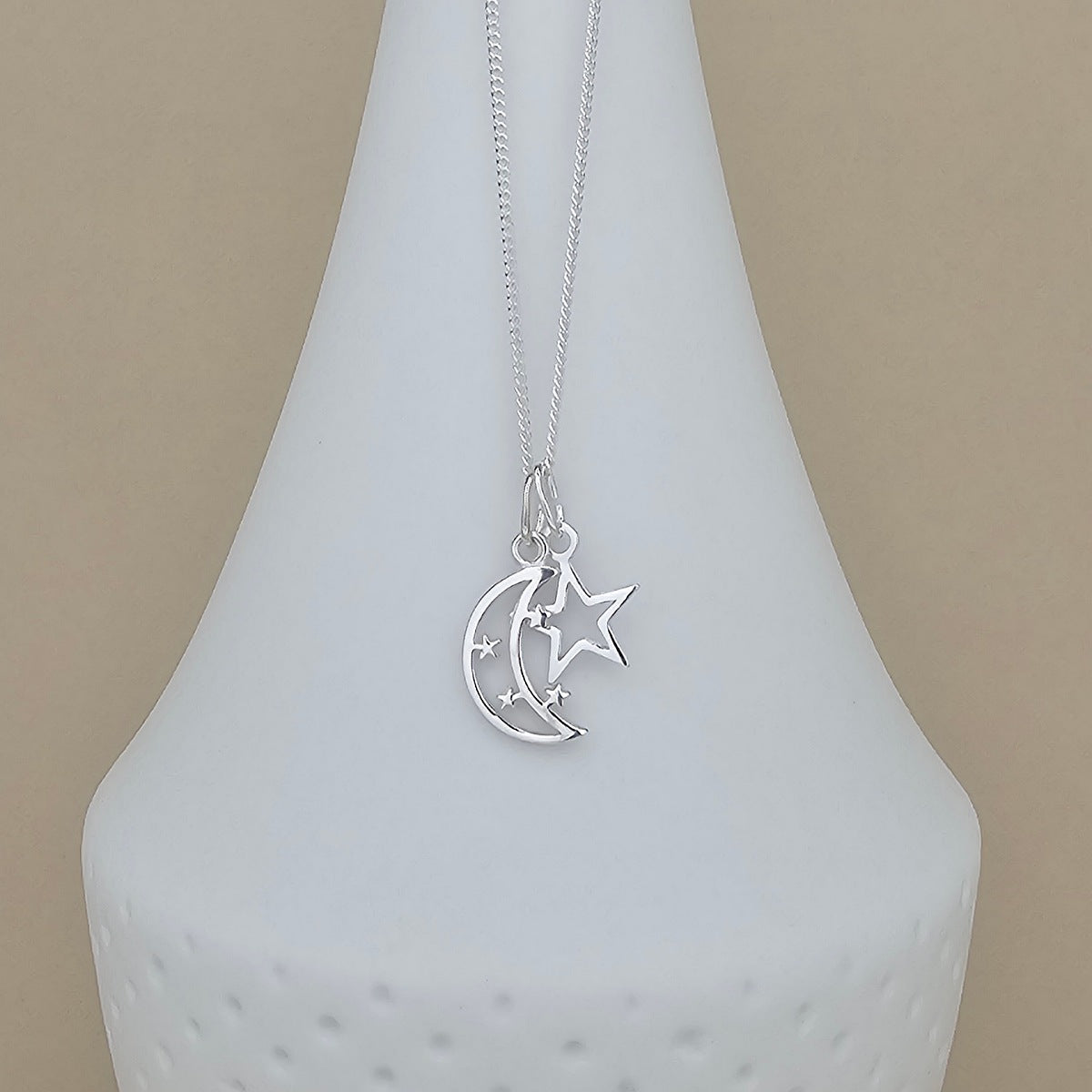 A silver necklace with a crescent moon and star pendant, displayed on a white ceramic vase against a beige background.