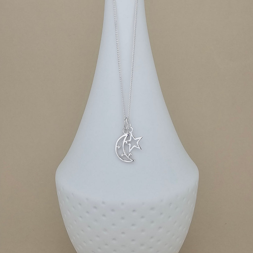 A silver crescent moon and star pendant necklace displayed on a white ceramic vase against a warm beige background.