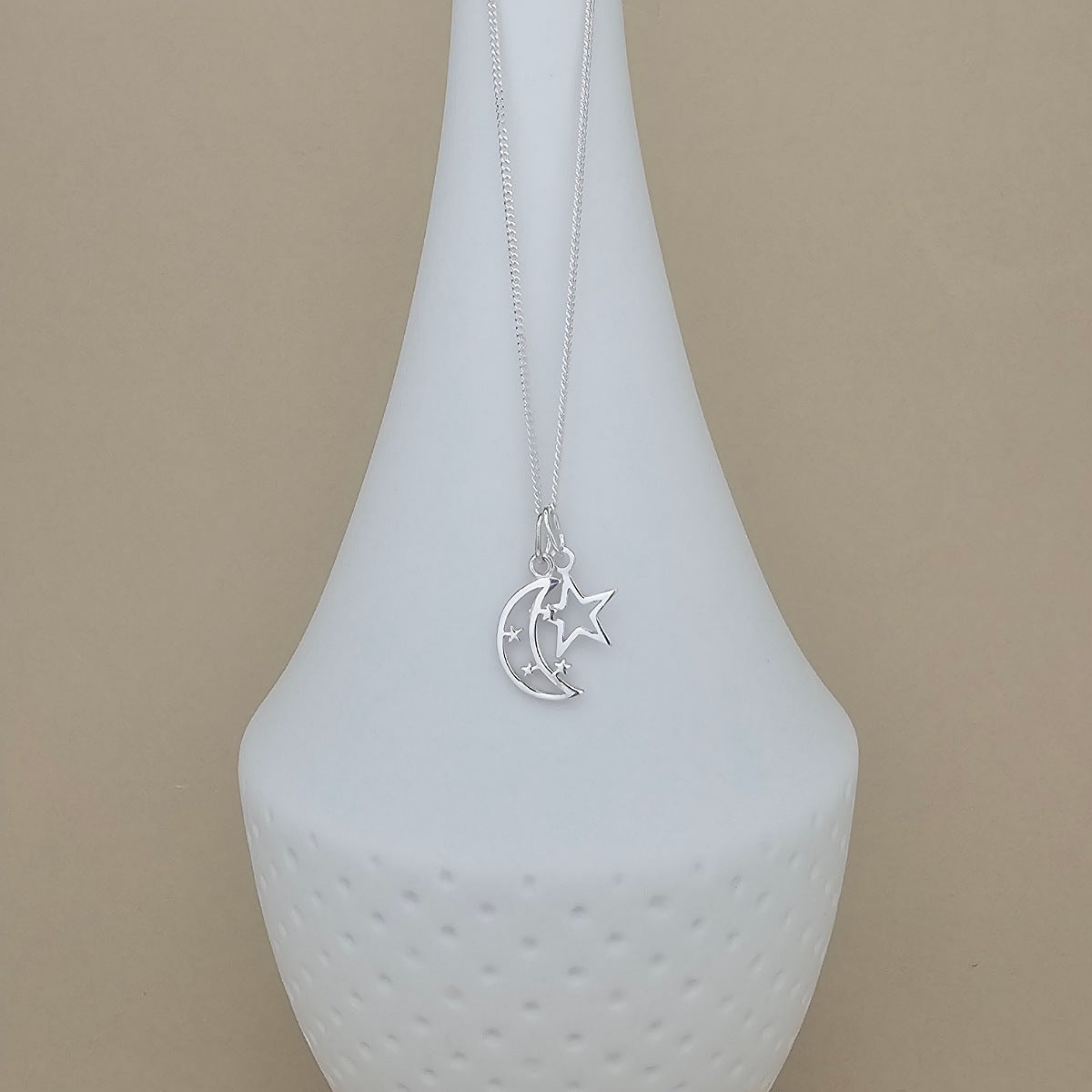 A silver crescent moon and star pendant necklace displayed on a white ceramic vase against a warm beige background.