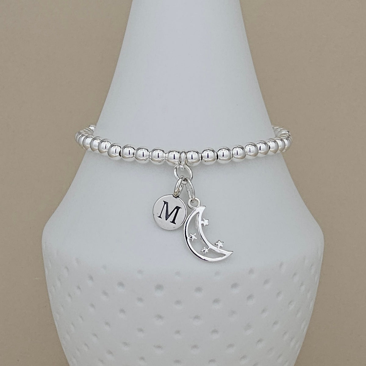 A silver beaded bracelet with a round initial 'M' charm and a crescent moon charm, displayed on a white textured cone stand against a beige background.