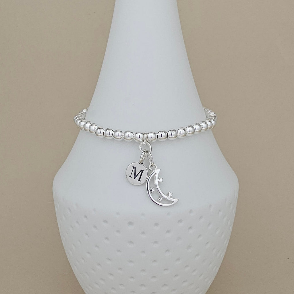 A silver beaded bracelet with a crescent moon and star charm, and a round initial 'M' charm, displayed on a white ceramic cone against a beige background.