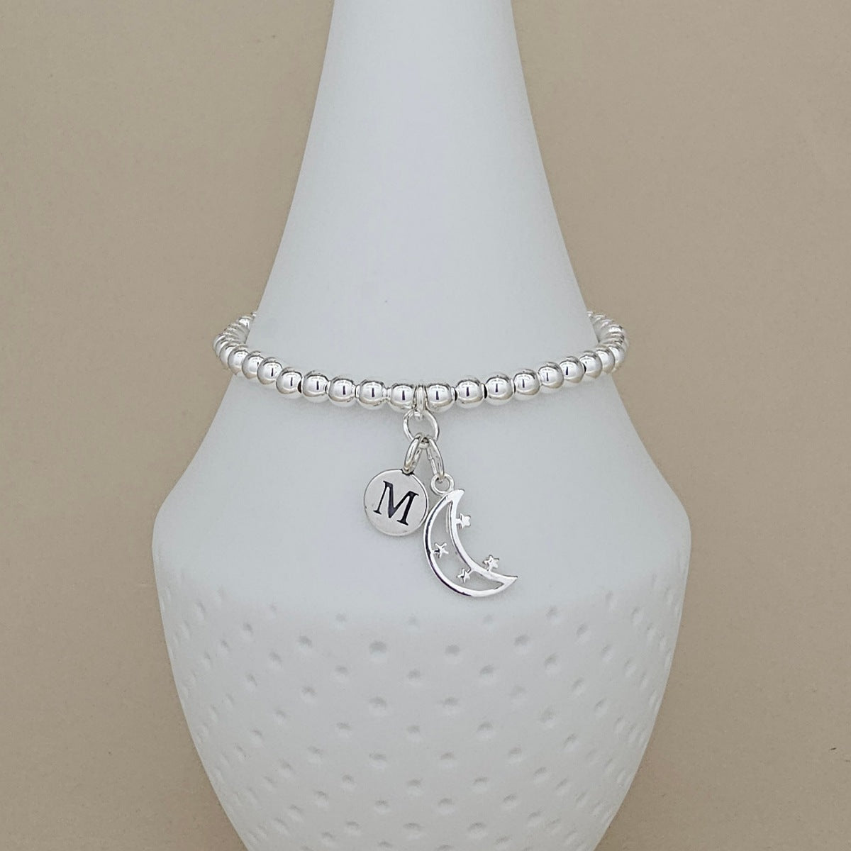 A silver beaded bracelet with a crescent moon and star charm, and a round initial 'M' charm, displayed on a white ceramic cone against a beige background.