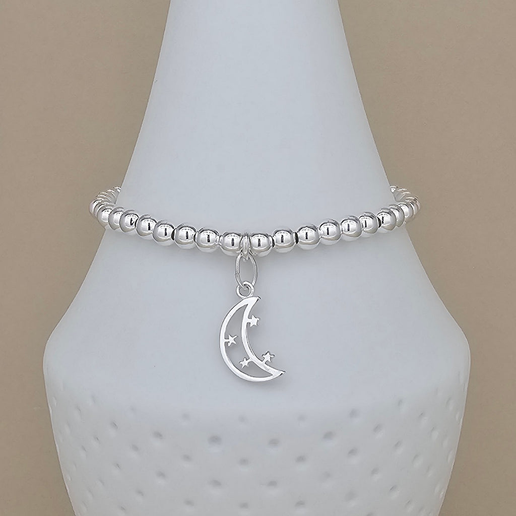 A silver beaded bracelet with a crescent moon and star charm, displayed on a white ceramic jewelry stand against a beige background.