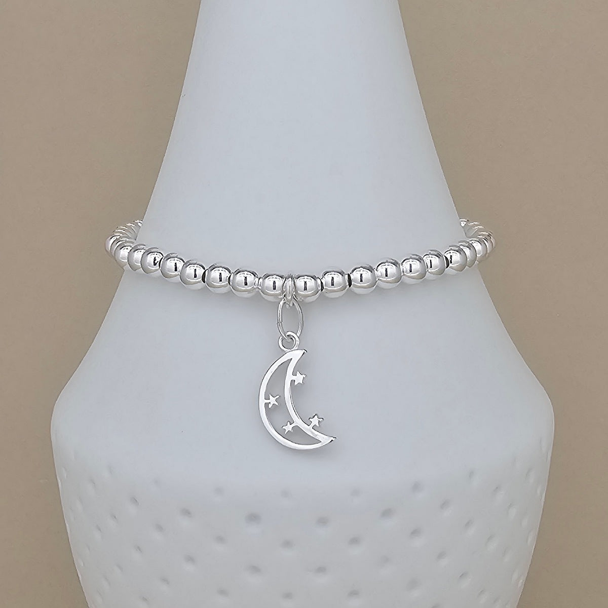 A silver beaded bracelet with a crescent moon and star charm, displayed on a white ceramic jewelry stand against a beige background.