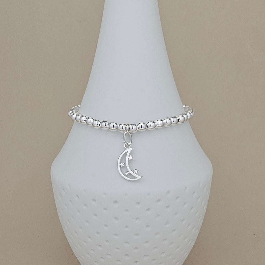A silver beaded bracelet with a crescent moon and star charm, displayed on a white ceramic jewelry cone against a beige background.