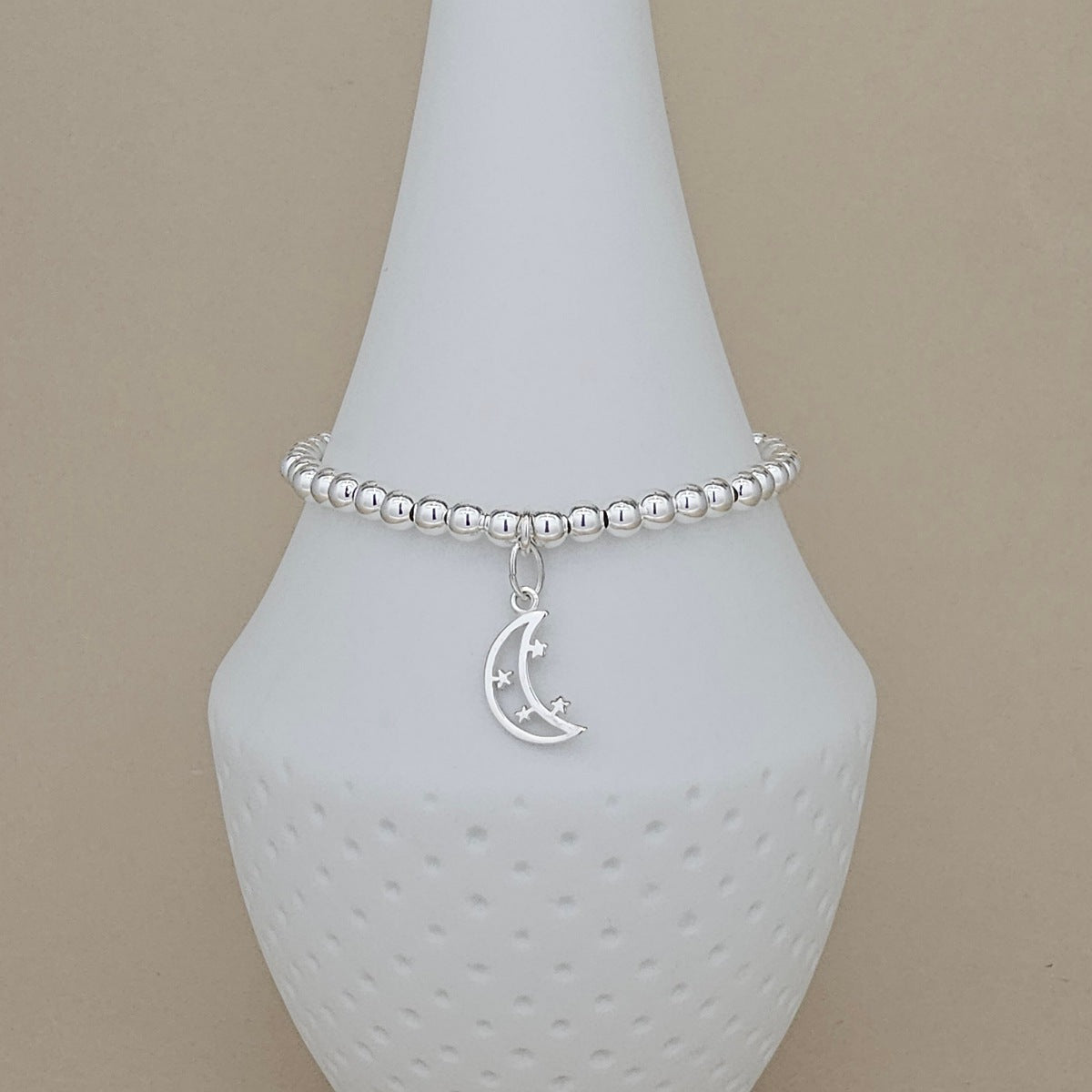 A silver beaded bracelet with a crescent moon and star charm, displayed on a white ceramic jewelry cone against a beige background.