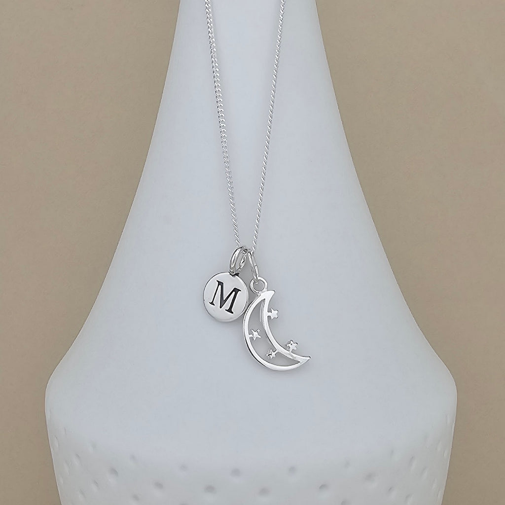 A silver necklace with a delicate chain and two charms: a round disc engraved with the letter 'M' and a crescent moon with small star details, displayed on a white ceramic vase against a beige background.