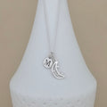 A silver necklace with a delicate chain and two charms: a round disc engraved with the letter 'M' and a crescent moon with small star details, displayed on a white ceramic vase against a beige background.