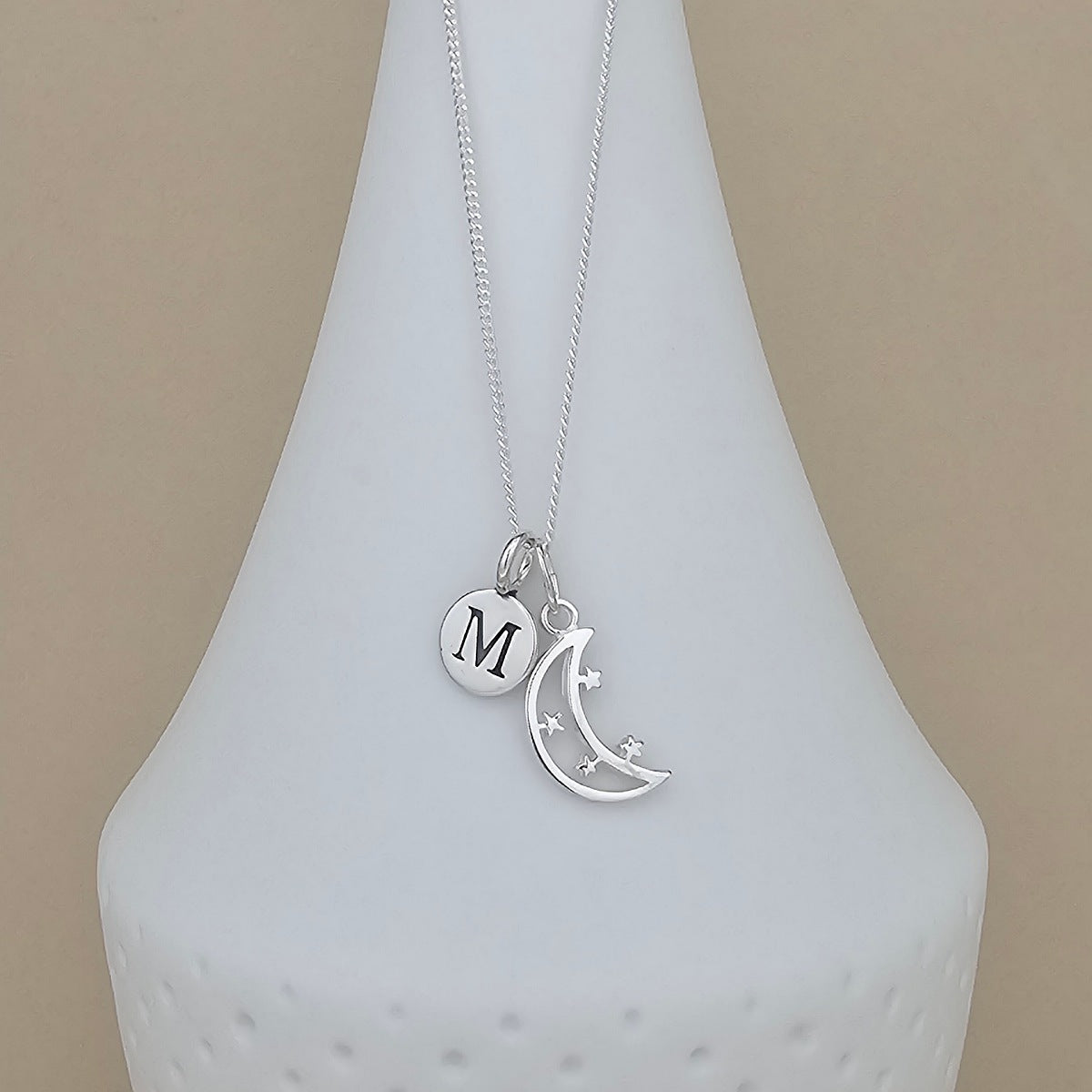 A silver necklace with a delicate chain and two charms: a round disc engraved with the letter 'M' and a crescent moon with small star details, displayed on a white ceramic vase against a beige background.