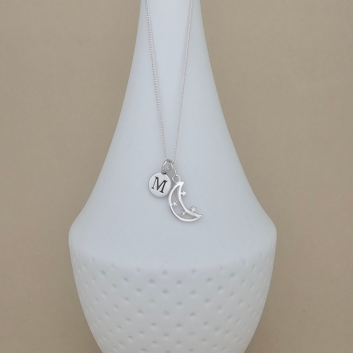 A silver necklace with a crescent moon and star pendant, alongside a small round initial 'M' charm, displayed on a white textured vase against a beige background.