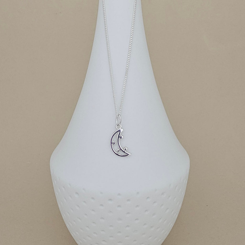 A silver crescent moon pendant necklace with small star details, displayed on a white ceramic vase against a beige background.