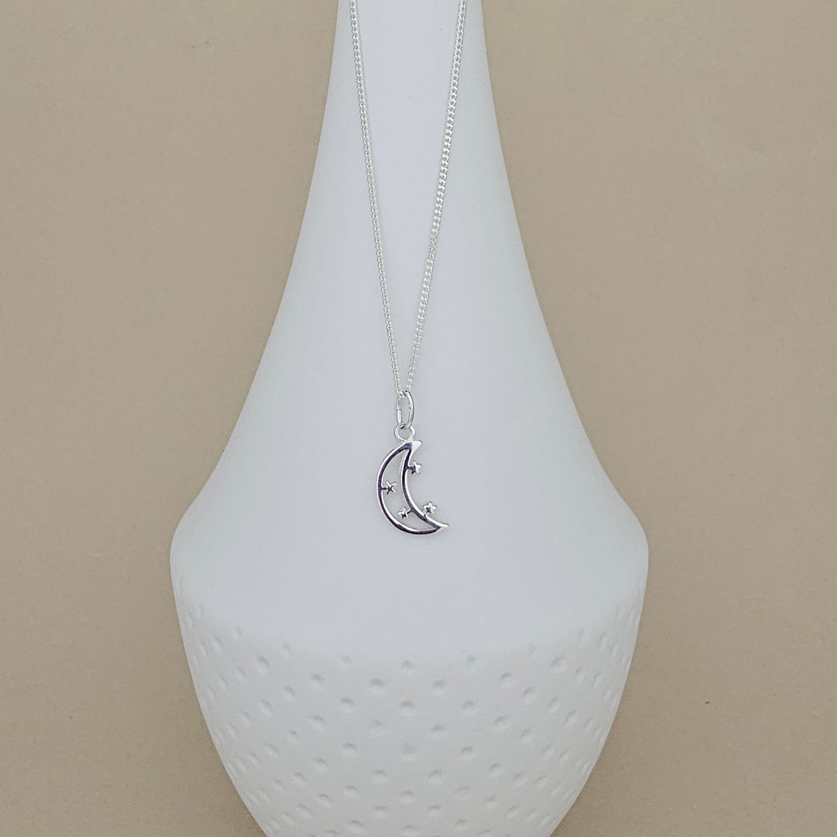 A silver crescent moon pendant necklace with small star details, displayed on a white ceramic vase against a beige background.