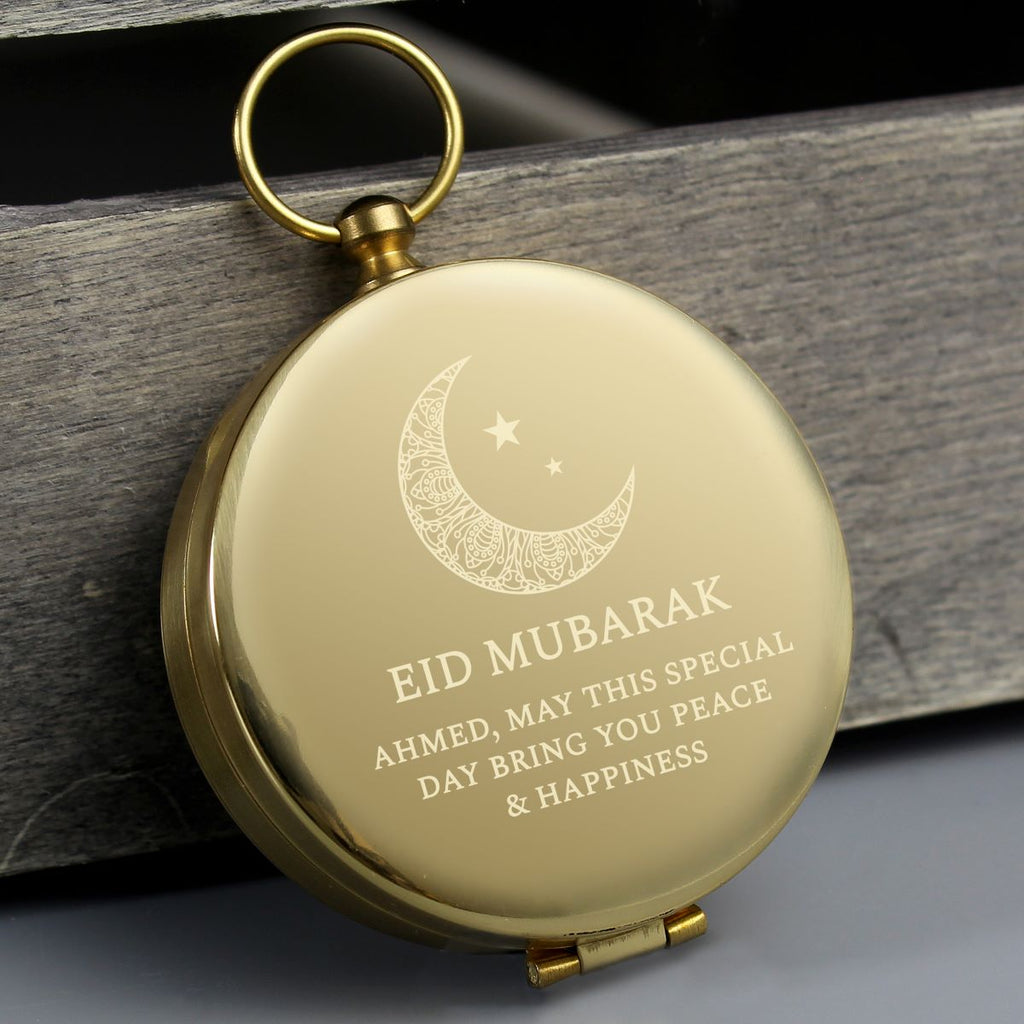 A round gold-toned metal compass with a hanging loop, shown closed on a dark wooden surface. The front is engraved with a crescent moon, stars, and the text 'EID MUBARAK AHMED, MAY THIS SPECIAL DAY BRING YOU PEACE & HAPPINESS'.