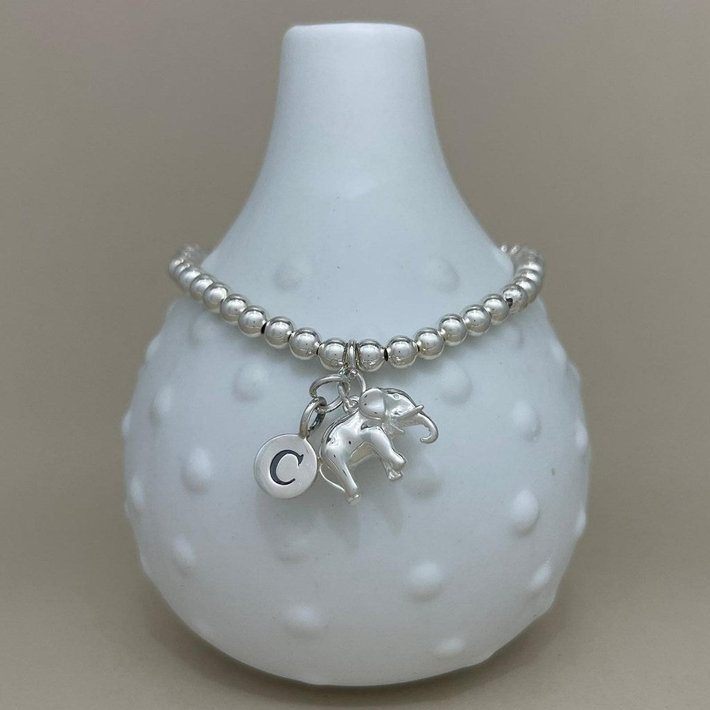 A silver beaded bracelet with a small elephant charm, a letter C initial charm, and a round charm, displayed on a white textured ceramic vase against a neutral beige background.