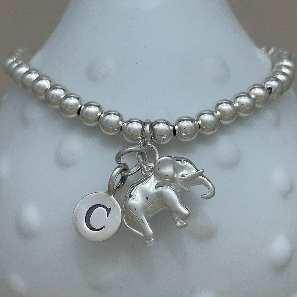 A silver beaded bracelet displayed on a white textured bust, featuring a dangling elephant charm and a round initial 'C' charm.