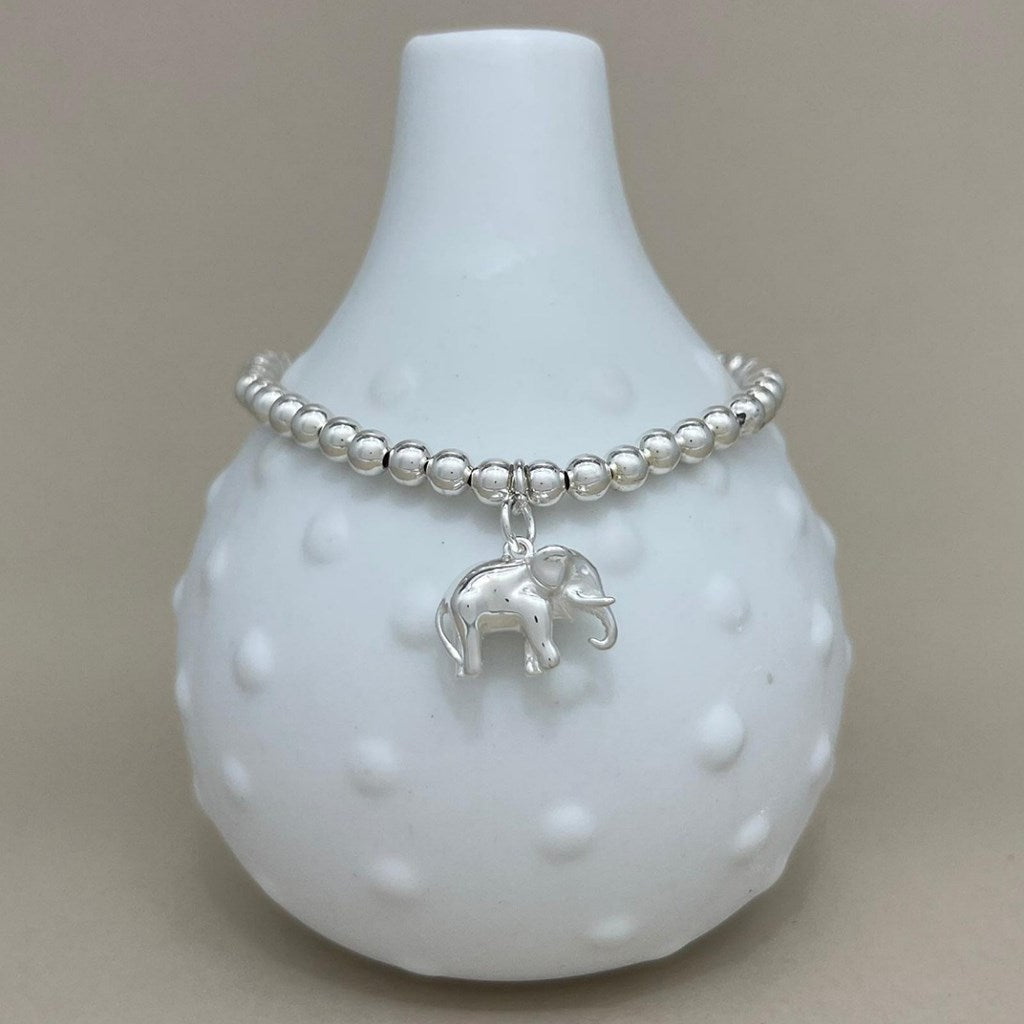 A silver beaded bracelet with a small elephant charm, displayed on a white textured glass vase against a neutral beige background.