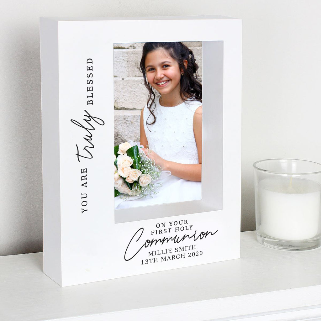 A white personalized Communion photo frame with black script text, shown on a white shelf next to a glass candle. The frame features a photo of a young girl in a white dress holding a bouquet, with custom text reading 'YOU ARE Truly BLESSED' and 'ON YOUR FIRST HOLY Communion MILLIE SMITH 13TH MARCH 2020'.