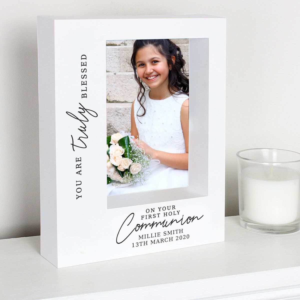 A white personalized Communion photo frame with black script text, shown on a white shelf next to a glass candle. The frame features a photo of a young girl in a white dress holding a bouquet, with custom text reading 'YOU ARE Truly BLESSED' and 'ON YOUR FIRST HOLY Communion MILLIE SMITH 13TH MARCH 2020'.