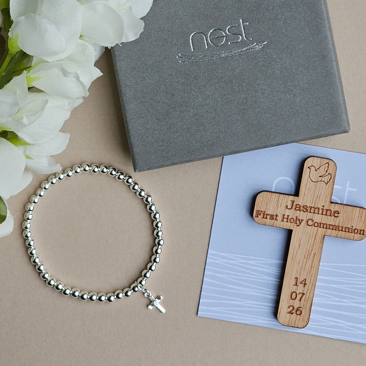 A silver beaded bracelet with a small cross charm is displayed next to a personalized wooden cross engraved with 'Jasmine First Holy Communion' and dates '14 07 26', alongside a gray Nest gift box and a light blue card on a beige background with white flowers.