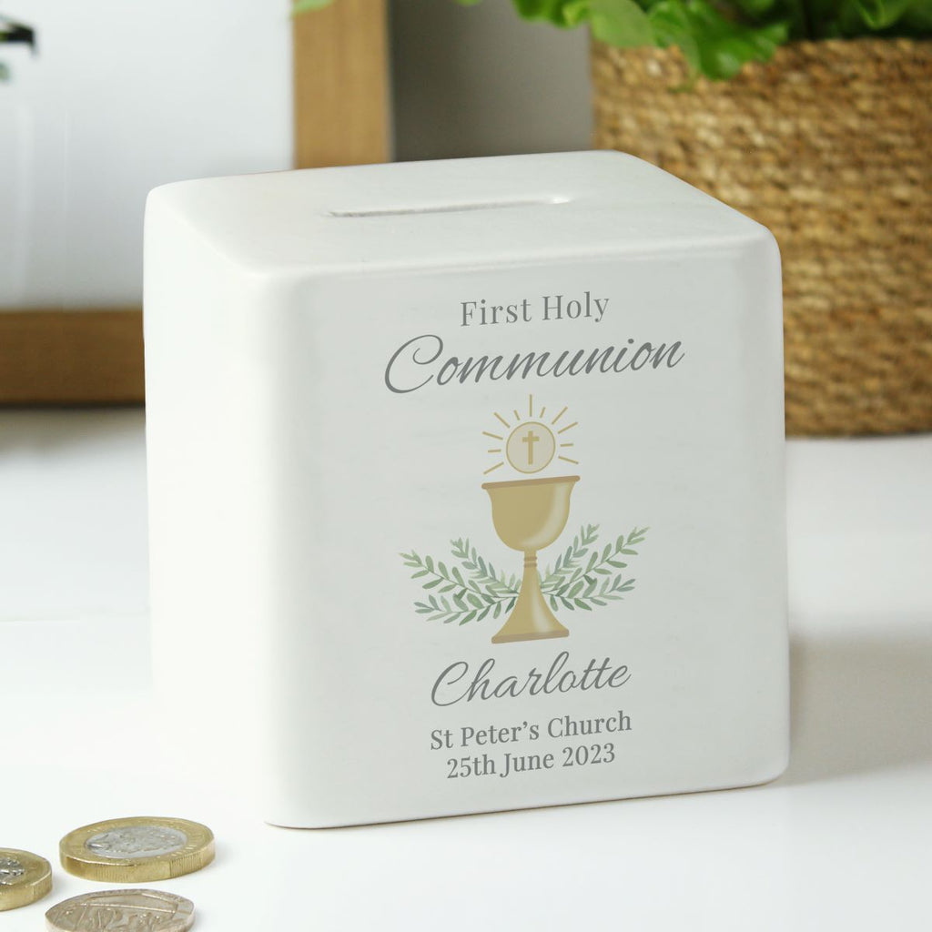 A white ceramic First Holy Communion money box with a personalized design, featuring a gold chalice illustration and the name 'Charlotte' on a white background with coins in the foreground.